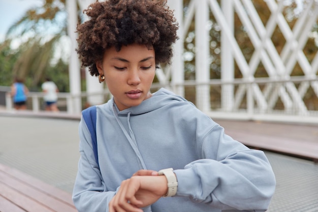 Tiro ao ar livre de mulher milenar séria com cabelo encaracolado verifica o tempo no relógio de pulso espera por alguém vestido com capuz azul posa contra o fundo desfocado durante o dia conceito de estilo de vida