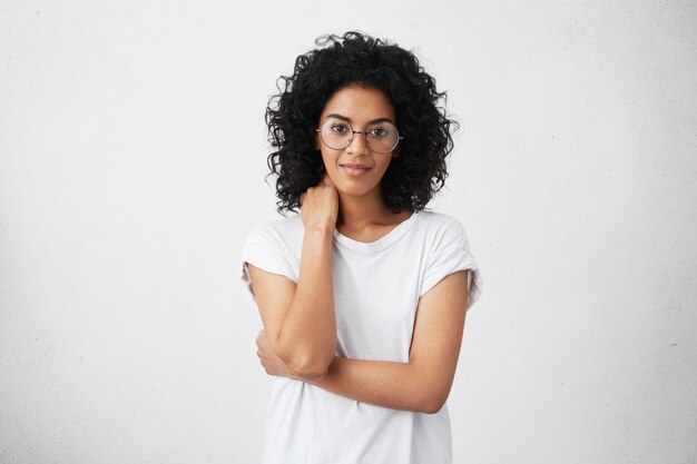 Tiro colhido da mulher afro-americana nova sorridente fofa amigável bonita posando com postura fechada, ligeiramente sorrindo, tendo olhar tímido, usando óculos. Emoções e gestos humanos