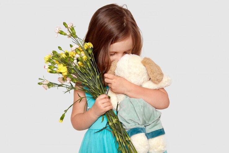 Foto grátis tiro de adorável criança do sexo feminino brinca com seu brinquedo favorito, detém flores, gosta de receber presente, vestido com roupas festivas, isoladas no branco. crianças e estilo de vida