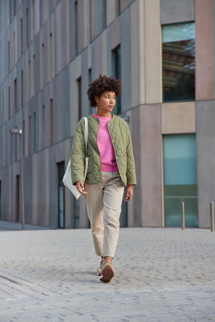 Tiro de corpo inteiro de mulher de cabelos cacheados usa calças de jaqueta e tênis carrega saco de tecido passeios ao ar livre no centro da cidade perto do edifício moderno chega ao destino necessário. Estilo de vida urbano