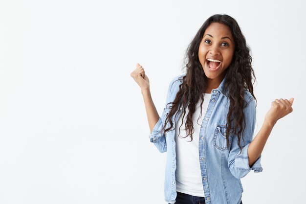 tiro de mulher bem sucedida de pele escura, com longos cabelos ondulados, vestindo camisa jeans apertando os punhos de emoção, sendo feliz em comemorar sua conquista e sucesso.