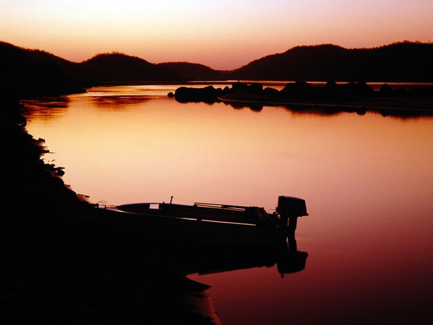 Tiro de silhueta de uma lancha no corpo de um lago cercado por montanhas durante o pôr do sol