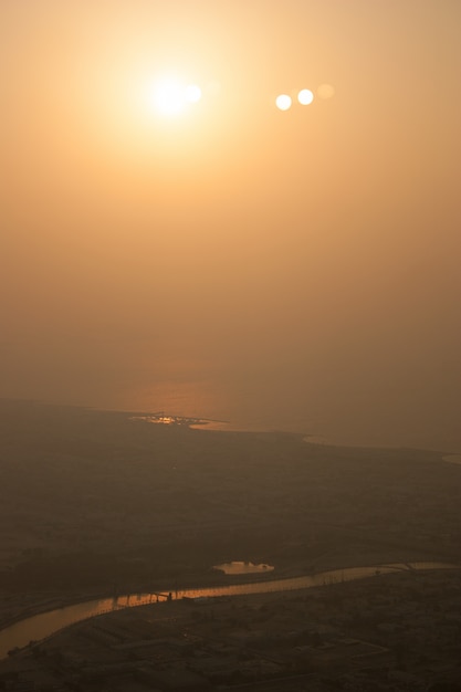 Tiro distante de um rio que flui perto de uma cidade durante o dia com um sol brilhando no céu