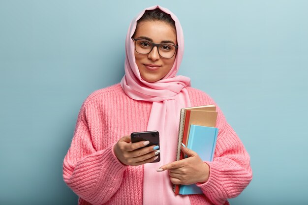 Tiro horizontal de estudante universitário satisfeito usa novo aplicativo legal no celular, carrega bloco de notas para escrever notas, usa óculos, lenço de seda e suéter de tricô, isolado sobre a parede azul.