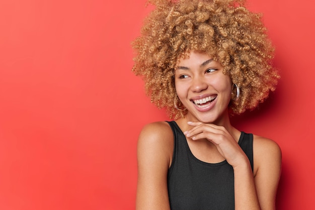 Tiro horizontal de jovem feliz com cabelo loiro encaracolado mantém a mão sob o queixo sorri agradavelmente vestido com camiseta preta isolada sobre o espaço de cópia de fundo vermelho para o seu conteúdo promocional