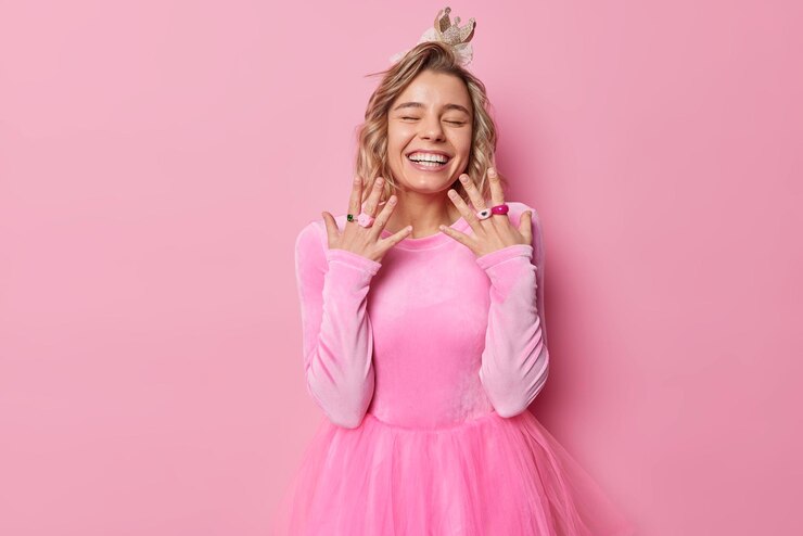 Foto grátis tiro horizontal de jovem feliz usa vestido festivo e coroa mantém as mãos levantadas os olhos fechados estando de bom humor posa contra o fundo rosa se prepara para o conceito de férias de festa a fantasia