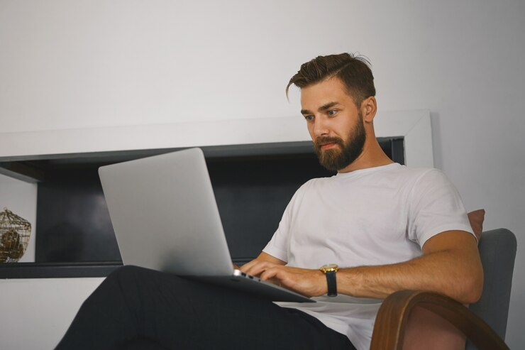 Foto grátis tiro horizontal de jovem freelancer masculino bonito com barba espessa, sentado na poltrona com o computador portátil genérico, trabalhando remotamente de casa. conceito de pessoas, dispositivos, tecnologia e comunicação