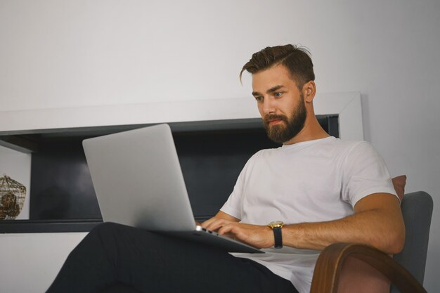 Tiro horizontal de jovem freelancer masculino bonito com barba espessa, sentado na poltrona com o computador portátil genérico, trabalhando remotamente de casa. Conceito de pessoas, dispositivos, tecnologia e comunicação