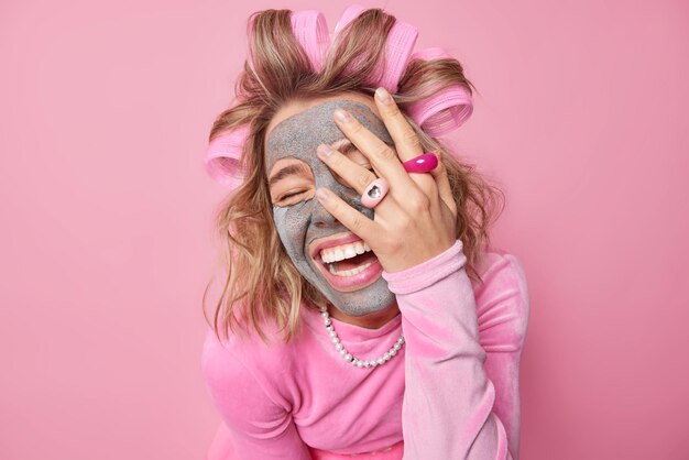 Tiro horizontal de jovem muito feliz cobre o rosto com a palma da mão aplica máscara de argila de beleza no rosto para remover linhas finas faz com que o penteado queira ficar bonito em poses de ocasiões especiais no interior