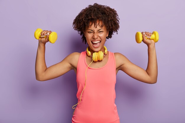 Tiro horizontal de mulher Afro feliz energizada levanta os braços com halteres, gosta de treinamento esportivo com música em fones de ouvido, vestida com colete rosa casual, poses internas. Pessoas