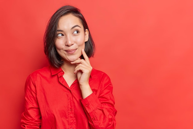 Tiro horizontal de mulher bonita com cabelo escuro mantém o dedo na bochecha desvia o olhar tem expressão sonhadora usa camisa isolada sobre fundo vermelho espaço de cópia em branco para o seu conteúdo promocional