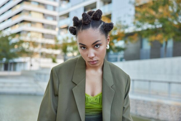 Tiro horizontal de mulher bonita vestida com jaqueta verde escura elegante tem penteado de coque parece autoconfiante em poses de câmera ao ar livre na cidade contra fundo desfocado Conceito de estilo de vida