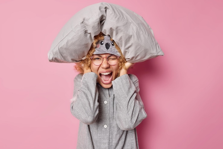 Foto grátis tiro horizontal de mulher emocional exclama em voz alta não pode adormecer por causa do ruído mantém travesseiro macio sobre a cabeça cobre as orelhas vestidas com roupa de dormir isolada sobre fundo rosa interrupção do sono