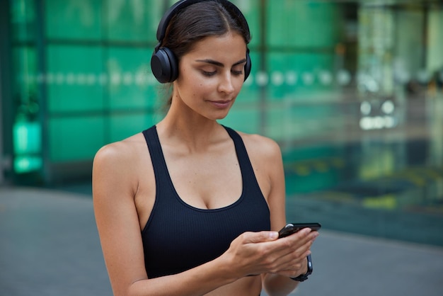 Tiro horizontal de mulher esportiva verifica o celular antes de fazer alguns exercícios ouve música da lista de reprodução através de fones de ouvido sem fio vestidos de top preto caminha fora contra fundo desfocado