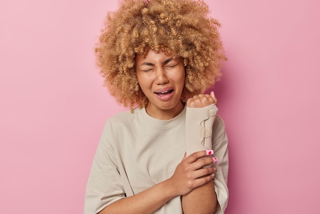 Tiro horizontal de mulher infeliz com cabelos cacheados chora de desespero sente dor depois de machucar o braço usa curativo enrolado no braço vestido com camiseta casual isolada sobre fundo rosa pessoas e lesões