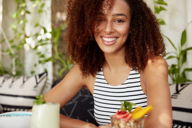 Tiro horizontal de mulher jovem alegre de pele escura com cabelo encaracolado, aproveita o tempo livre no fim de semana no café, come um delicioso coquetel. Mulher afro-americana feliz por passar as férias com amigos