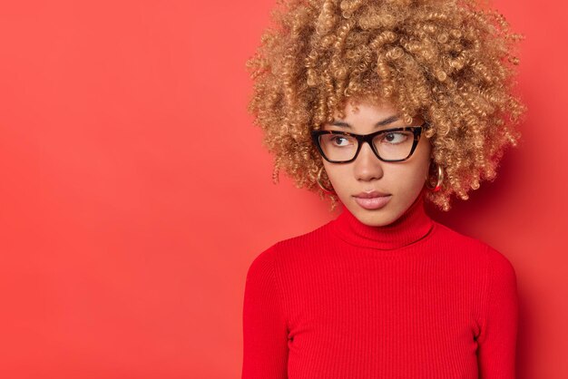 Tiro horizontal de mulher pensativa com cabelos cacheados focados em pensamentos profundos usa óculos ópticos poloneck casual isolado sobre o espaço de cópia de fundo vermelho para o seu conteúdo promocional