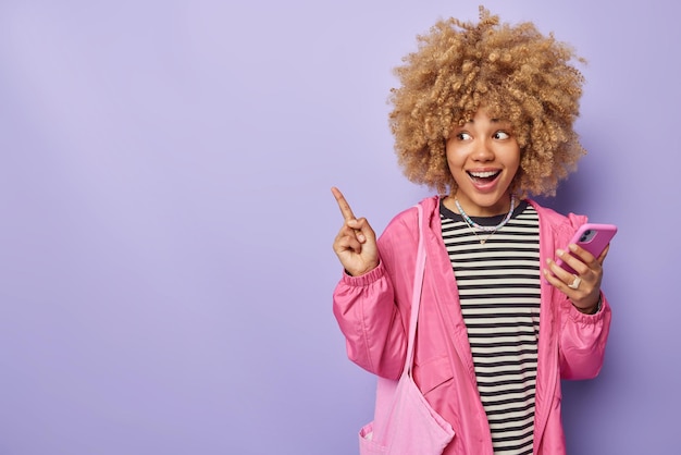 Tiro horizontal de mulher positiva com pontos de cabelo espesso encaracolado deixados no espaço em branco mostra o local de direção para o seu conteúdo de publicidade detém o telefone celular vestido com um jumper listrado e jaqueta