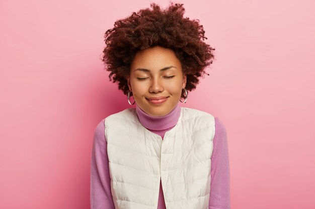 Tiro horizontal de mulher satisfeita tem penteado encaracolado, olhos fechados, expressão calma, usa brincos redondos e roupa casual, posa contra um fundo rosa.
