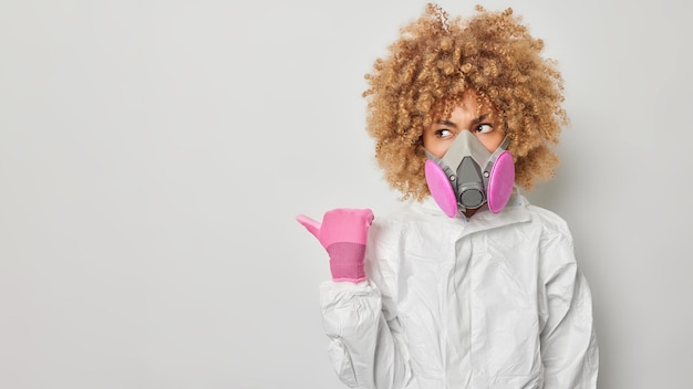 Foto grátis tiro horizontal de mulher séria com cabelos cacheados usa traje de proteção e respirador aponta o polegar para longe promove algo que diz sobre proteção contra radiação isolada sobre fundo cinza