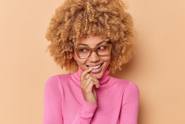 Tiro horizontal de mulher sonhadora feliz mantém o dedo indicador nos lábios parece alegremente afastado lembra boas memórias usa óculos e gola rosa isolada sobre fundo bege. Emoções positivas