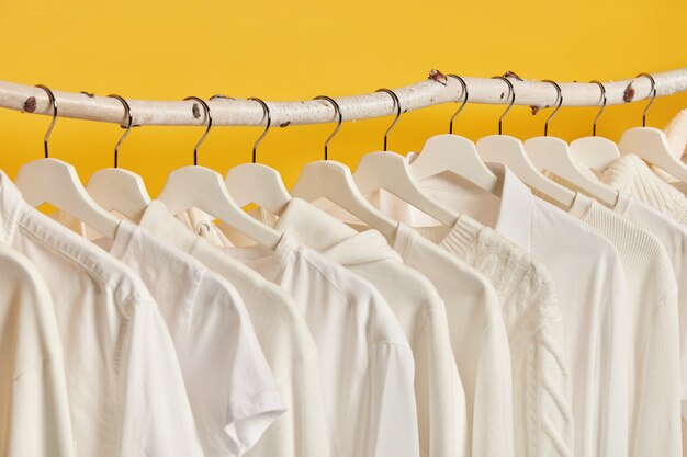 Tiro horizontal de roupas femininas brancas penduradas em racks, isolado sobre fundo amarelo. Camarim com roupas femininas.