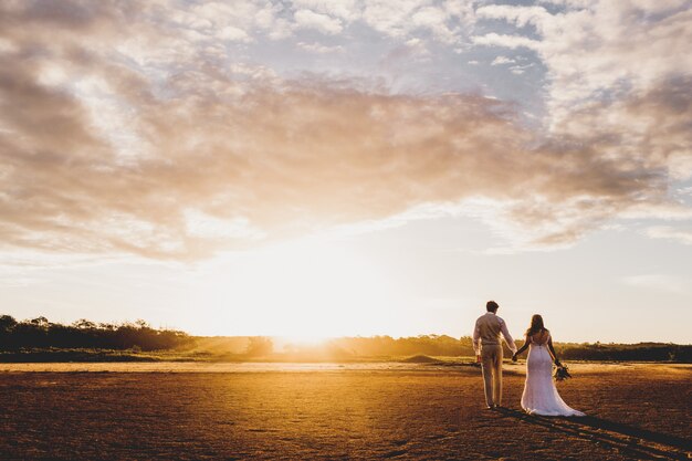 Tiro horizontal de um homem e uma mulher em trajes de casamento, de mãos dadas durante o pôr do sol
