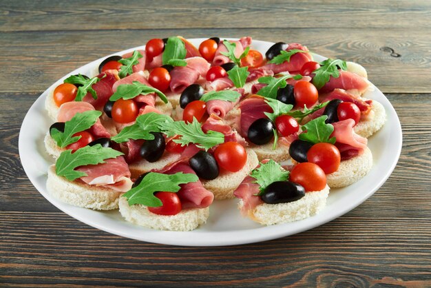 Tiro horizontal de um prato com canapés com presunto, tomate cereja e azeitonas pretas decorado com rucola rucoli planta vegetais comestíveis bacon jamon menu aperitivos restaurante.