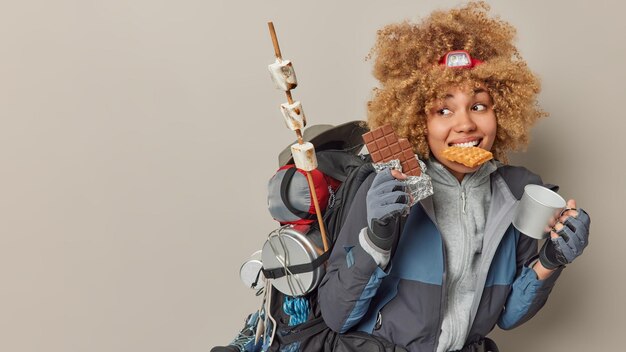 Tiro interior de alpinista de mulher alegre come waffles caseiros barra de chocolate e bebe chá após expedição carrega mochila se prepara para piquenique isolado sobre fundo cinza espaço em branco à parte