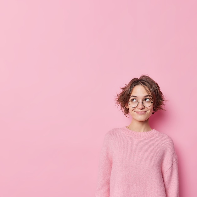 Foto grátis tiro interior de linda mulher sonhadora com cabelo escuro curto sorri suavemente e desvia o olhar usa óculos redondos e suéter confortável isolado sobre fundo rosa espaço de cópia em branco para sua promoção