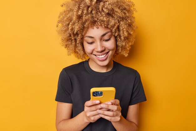 Foto grátis tiro interior de mulher bonita com cabelo encaracolado usa tipos de telefone celular mensagem sms sorri alegremente vestido com camiseta preta casual isolada sobre fundo amarelo conceito de tecnologias modernas