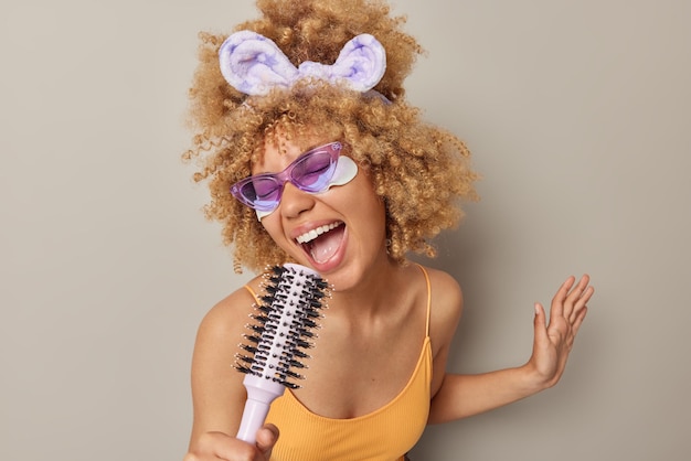 Foto grátis tiro interior de mulher loira alegre segura pente de cabelo perto da boca como se o microfone se sentisse muito feliz aplica manchas de beleza sob os olhos usa óculos de sol roxos faixa de cabeça e camiseta isolada na parede cinza