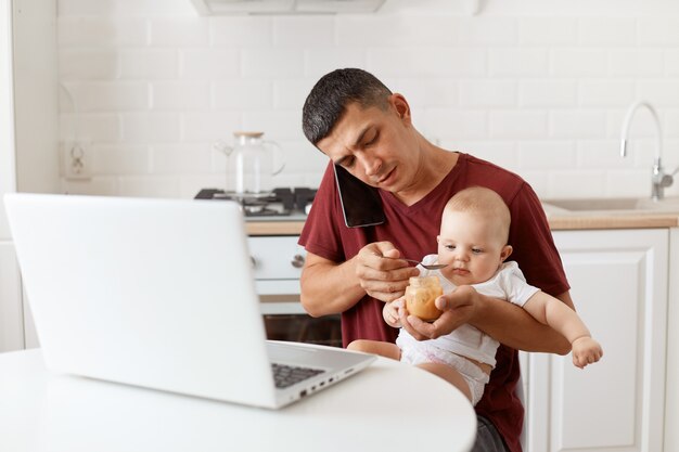 Tiro interno de homem moreno vestindo camiseta marrom estilo casual t, sentado à mesa na cozinha, falando via smartphone, alimentando sua filha com purê de frutas.