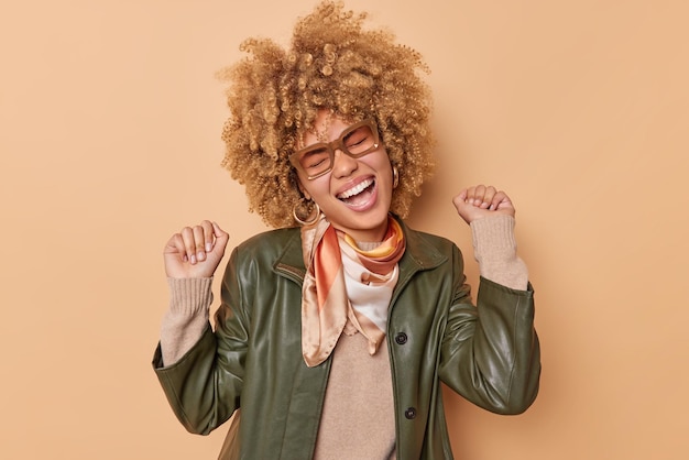 Foto grátis tiro interno de jovem despreocupada otimista sacode danças de punhos e se sente bem vestido com um lenço de jaqueta de couro ao redor do pescoço se sente muito feliz isolado sobre um fundo bege gosta de música.