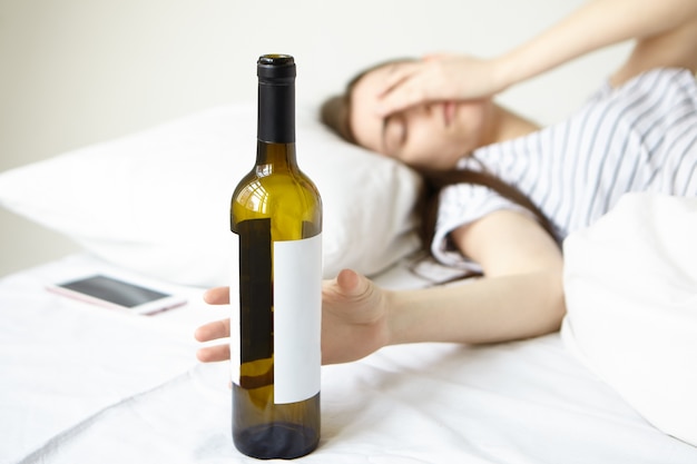 Foto grátis tiro interno de jovem morena deitada na cama branca, tocando o rosto, tendo uma forte dor de cabeça depois da festa à noite, acordando de manhã com ressaca. foco seletivo na mão segurando uma garrafa de vinho