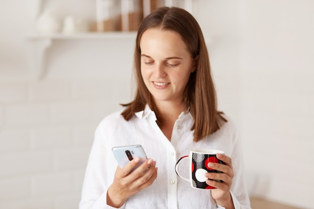 Tiro interno de mulher bonita adulta jovem usando telefone celular e tomando um café na cozinha, segurando a xícara de bebida nas mãos e olhando para o visor do dispositivo.