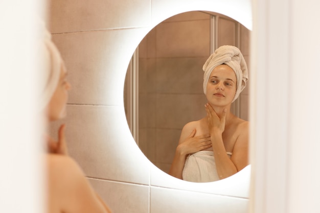 Tiro interno de mulher bonita olhando no reflexo no espelho depois do banho, tocando seu pescoço, fazendo procedimentos matinais, apreciando seu olhar fresco.