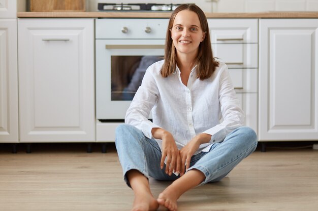 Tiro interno de mulher sorridente com cabelo escuro, vestindo calça jeans e camisa branca, sentada no chão em uma sala iluminada contra o conjunto de cozinha, olhando para a câmera com uma expressão positiva.