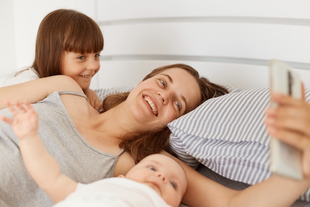 Tiro interno de mulher sorridente feliz vestindo camiseta sem mangas cinza, deitada na cama com seus filhos e segurando o smartphone nas mãos, tendo a videochamada com o marido.