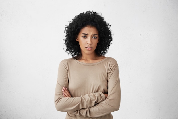 Tiro interno de uma jovem cética de raça mista que se sente desconfiada, seu olhar expressando desaprovação ou dúvida, mantendo os braços cruzados enquanto suspeita que o marido a traiu. Horizontal