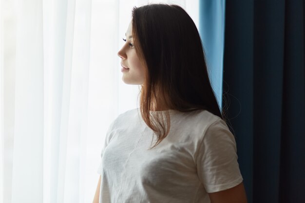 Tiro lateral da mulher de cabelos escuros pensativo vestido com camiseta branca, pensa em algo enquanto fica perto da janela