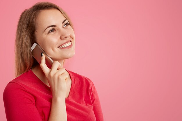 Tiro lateral de atraente mulher feliz tem expressão sonhadora, fala via telefone celular, gosta de conversar com um amigo, usa camisola vermelha