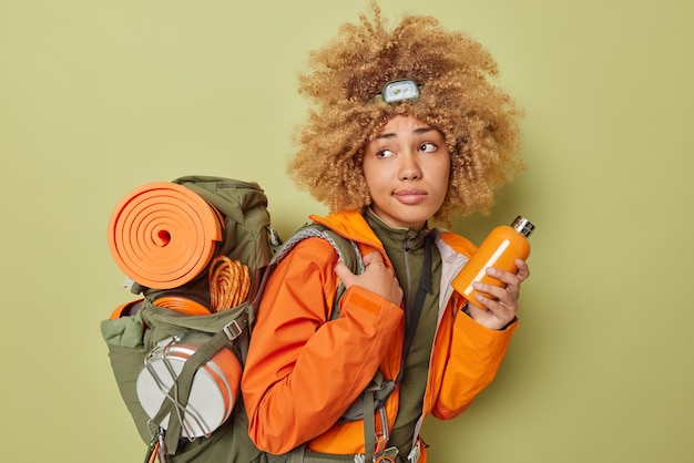 Tiro lateral de campista de mulher pensativa de cabelos encaracolados segura garrafa com água fresca vestida com jaqueta laranja carrega poses de mochila contra fundo verde escuro conceito de caminhada e turismo