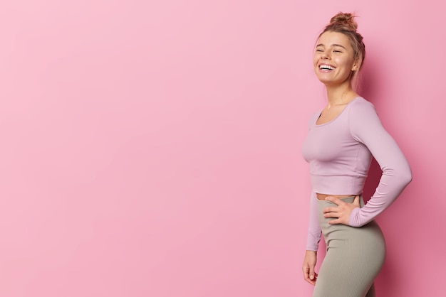 Tiro lateral de mulher magra saudável vestida com roupas esportivas ri alegremente tem um sorriso largo satisfeito após o exercício tem poses regulares de treinamento físico contra o espaço de cópia em branco de fundo rosa