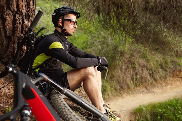 Tiro lateral do atraente feliz jovem ciclista europeu em equipamento de proteção, sentado sob uma árvore com seu veículo motorizado de duas rodas e contemplando a incrível natureza selvagem ao seu redor