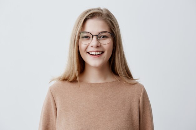 Tiro na cabeça da mulher caucasiana jovem de aparência agradável, usando óculos com sorriso largo, mostrando os dentes retos brancos sendo felizes por causa de notícias positivas. Menina loira com sorriso agradável