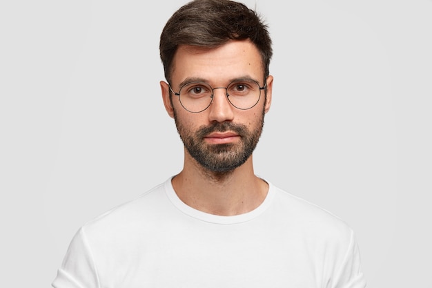 Tiro na cabeça de um freelancer masculino bonito com aparência atraente, tem barba e bigode escuros, parece diretamente com uma aparência séria, usa roupas casuais brancas. Monocromático. Expressões faciais.
