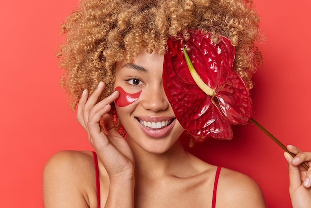 Tiro na cabeça de uma jovem bonita positiva usa produtos cosméticos orgânicos, aplica almofadas de colágeno e cobre os olhos com sorrisos de calla dentuçosamente isolados sobre um fundo vermelho vivo. conceito de beleza feminina