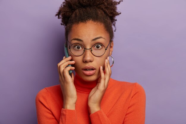 Tiro na cabeça de uma mulher de pele escura emocional atordoada engasga de medo, ouve notícias desagradáveis, tem uma conversa ao telefone, usa óculos ópticos e gola olímpica, modelos sobre fundo roxo.