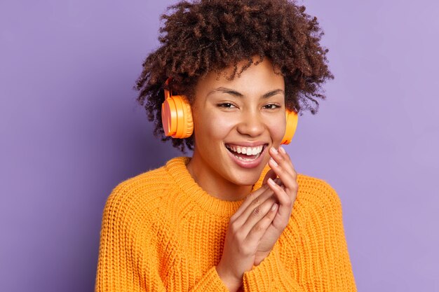 Tiro na cabeça de uma mulher de pele escura positiva ouve música em fones de ouvido sem fio estéreo, rindo e mantendo as mãos juntas usa um macacão casual